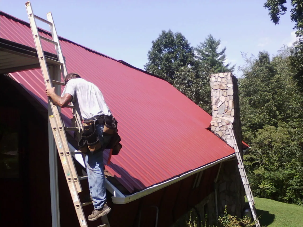 Roofer on a ladder beside a red metal roof and stone chimney — Soffit & Fascia Repair by Roofing Brothers in Shapleigh