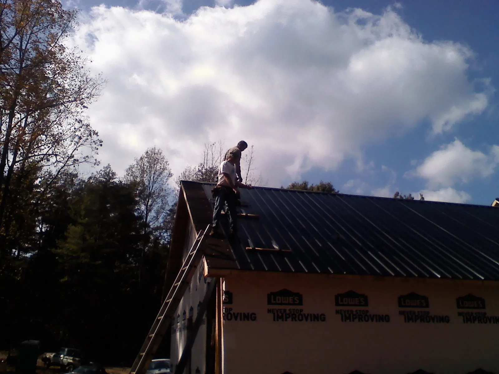 Roofer fastening dark metal panels high against a cloudy sky, from a Roof Inspection job in Shapleigh